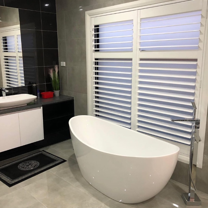 Freestanding bath beside plantation shutters and a dark tiled vanity